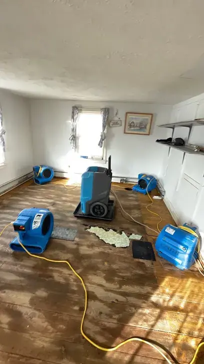 Air movers and a dehumidifier drying a water-stained wood floor in an emptied room